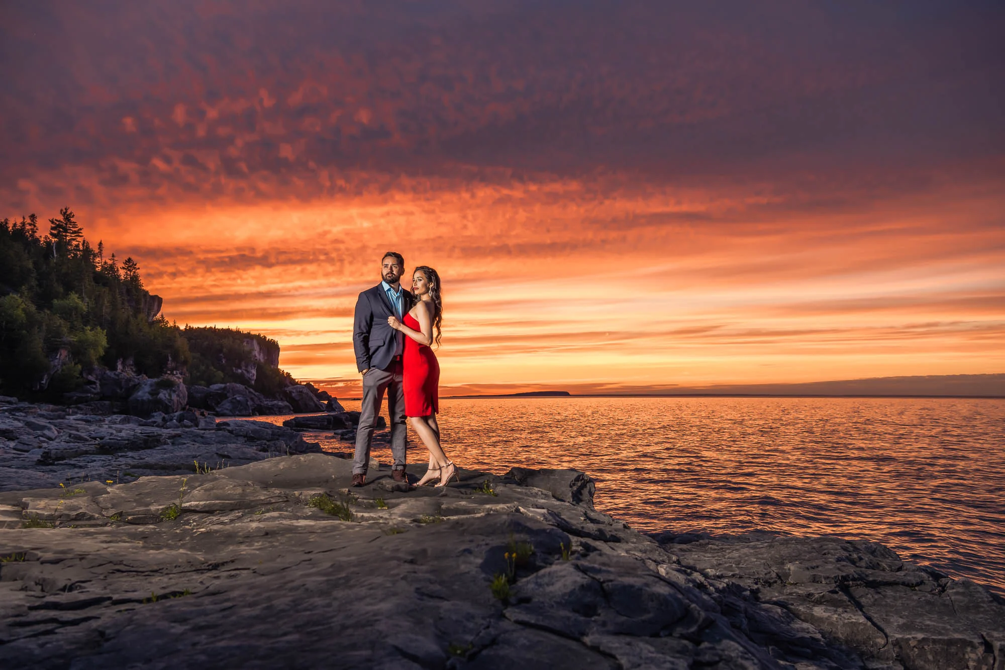 A couple on the rocky shore beneath a vibrant orange sunset